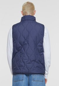 Navy quilted vest with a high collar, featuring wave-patterned stitching, worn over a white long-sleeve shirt and blue jeans.