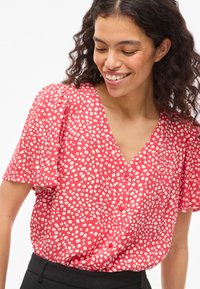 Femme souriante aux cheveux bouclés portant un chemisier rouge à manches courtes avec des pois blancs irréguliers et un pantalon noir, regardant vers le bas.