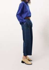 Pull bleu, pantalon large taille haute en denim foncé, ceinture marron, mocassins beiges à talon bloc. Matières texturées, coupe décontractée.