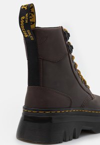 Bota de cuero marrón con una superficie texturizada, cordones amarillos y una gruesa suela de goma negra con un patrón acanalado. Incluye una lengüeta de tela negra.