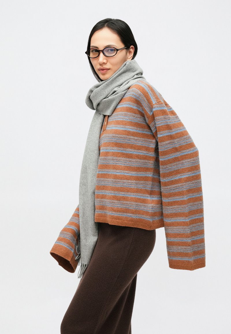 Maglione corto a righe marroni e grigie, tessuto morbido, abbinato a una sciarpa grigio chiaro. La modella indossa pantaloni scuri e occhiali rotondi.