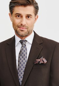 Brązowa marynarka w zestawieniu z białą koszulą, wzorzystym niebieskim krawatem oraz brązową chusteczką w kieszeni z kwiatowymi detalami.