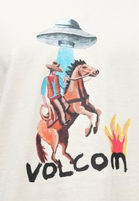 Λευκό πουκάμισο με γραφικό σχέδιο ενός καουμπόη πάνω σε άλογο, ενός UFO και φλόγες, με την επιγραφή «VOLCOM» με έντονα μαύρα γράμματα.