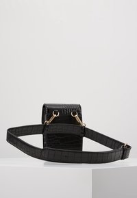 Svart konstläderväska med krokodilmönster, cylindrisk form, justerbar rem och detaljer i guld.