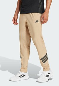 Beige sportieve broek met een taps toelopende pasvorm, voorzien van het zwarte Adidas-logo en gestreepte accenten; gecombineerd met witte sportschoenen.