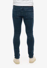 Mörkblå smal passform jeans med en slät denimtextur, med två bakfickor och subtila sömnadsdetaljer, som bärs med bruna skor.