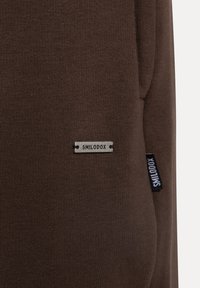 Nahaufnahme eines braunen Stoffes mit einem kleinen Metall- und Stoffetikett, auf dem der Markenname "SHILODOX" steht und das am Kleidungsstück befestigt ist.