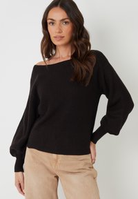 Zwarte geribde gebreide trui met een off-the-shoulder ontwerp en lange, losse mouwen, gecombineerd met lichtbeige broek. Textuur van de stof zichtbaar.