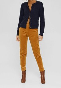 Cárdigan azul marino con cuello redondo y cierre de botones, combinado con pantalones de pana mostaza. El modelo lleva botas marrones hasta el tobillo.