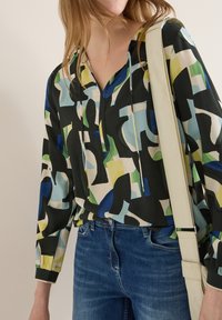 Femme portant un chemisier à motif abstrait multicolore et un jean bleu, avec une bandoulière de sac à épaule beige sur l'épaule.