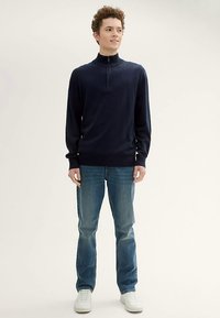 Temno modro pulover s patentnim zapiranjem, narejen iz mehkega materiala, z visokim ovratnikom in rebrastimi manšetami. Kombiniran s svetlo modrimi jeans hlačami in belimi supergami.