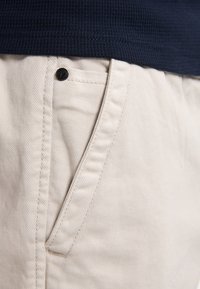 Beige bomulds chinos har en diagonalt ribbet tekstur, sidelommer og en sort knapdetalje på linningen. Marineblåt top er delvist synligt.