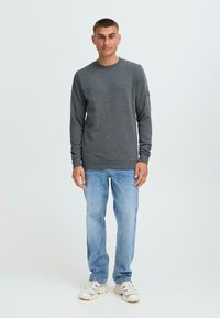 Maglione grigio con scollo rotondo, maniche lunghe e finitura testurizzata. Indossato con jeans in denim azzurro e sneakers bianche.