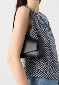 Černá syntetická kabelka s hladkým povrchem, jedním nastavitelným popruhem a klopovým uzávěrem, nošená na rameni osoby vedle vzorovaného topu.