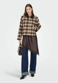 Kvinne står iført brun rutete jakke, asymmetrisk mørk brun skjørt over oppbrettede blå jeans, svarte hæler, holder en liten mørk veske.