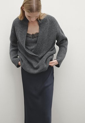 Cardigan en tricot gris avec un large col, présentant un motif texturé. Porté par-dessus une jupe en soie sombre avec une finition lisse.