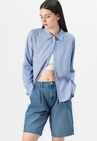 Camisa azul clara com riscas e botões, com colarinho, combinada com calções de ganga de cintura alta. Corte descontraído, bolsos frontais e detalhes plissados.