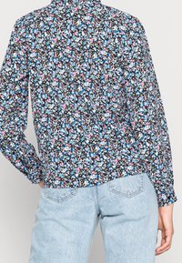 Camicia floreale nera, con un motivo di fiori blu e rosa, maniche lunghe e colletto, abbinata a jeans azzurri chiari.