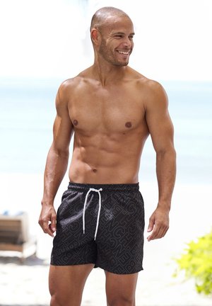 Lächelnder, oberkörperfreier Mann steht an einem Strand, trägt schwarze Badeshorts mit weißem Kordelzug und Textmuster.