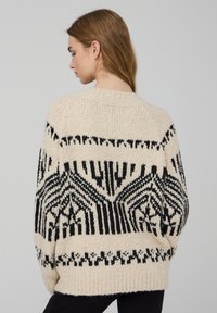 Maglione crema spesso con motivo geometrico nero, trama lavorata a maglia e polsini a coste. Presenta una vestibilità ampia e scollo rotondo.