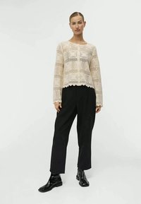 Pull en tricot crème avec un design en dentelle et un bord festonné, associé à un pantalon noir taille haute et des bottines noires brillantes.