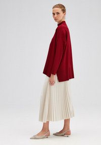 Burgundska pletena jopica z visokim ovratnikom, v kombinaciji z nagubanimi kremnimi midi krilom in bež slingback čevlji z dekorativnim okrasom.