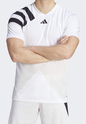 Homme en tenue de sport blanche avec des bandes diagonales noires sur l'épaule et le logo Adidas, bras croisés, torse visible, fond uni.