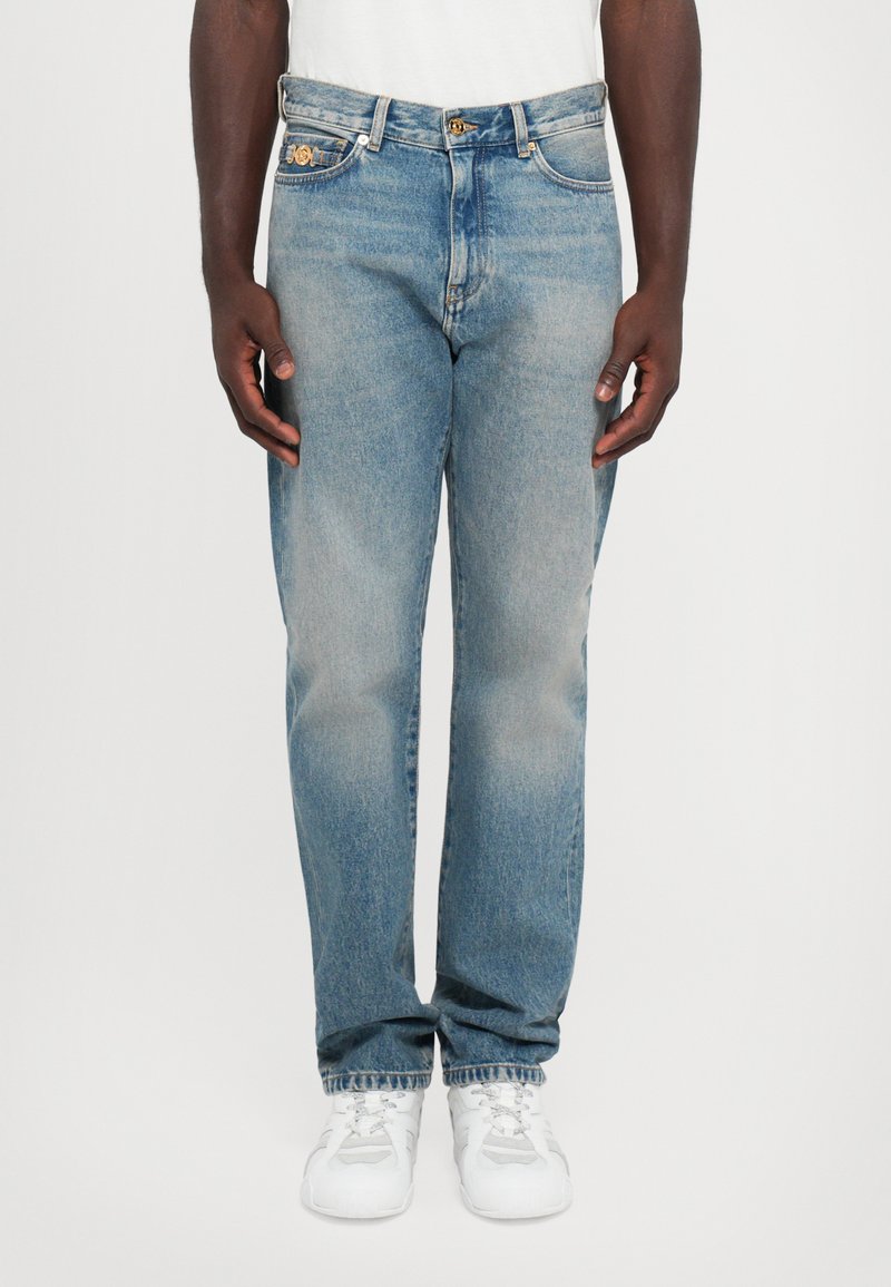 Man som bär blekta blåjeans och vita sneakers, står mot en enfärgad vit bakgrund med händerna avslappnat vid sidorna.