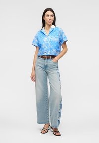 Mujer con camisa azul de manga corta con estampado, vaqueros de pierna ancha azul claro con diseños florales en los laterales, cinturón marrón y sandalias negras con tacón.