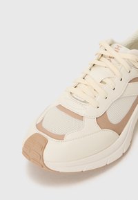 Sneaker bianco con una tomaia in pelle testurizzata e mesh, dettagli beige, punta tonda e design con lacci. Suola in gomma con trazione.