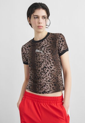 Ung person med kort, vådt hår iført leopardmønstret Puma T-shirt med sort kant og knaldrøde bukser, hænderne i lommerne, kigger mod venstre.