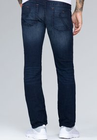 Jeans de mezclilla oscura con un corte recto, que cuentan con dos bolsillos traseros, costuras en contraste y áreas sutilmente desgastadas. Se llevan con zapatillas blancas.
