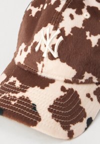 Brun og beige kuemønster lue med myk, teksturert stoff, med hvit brodert "NY"-logo og sydd detaljer langs skjermen.