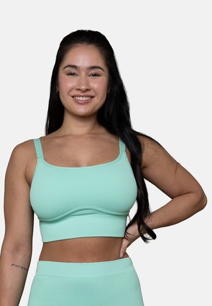 Frau mit langen schwarzen Haaren, die ein mintgrünes, geripptes Croptop und passende hochtaillierte Unterteile trägt, lächelt mit einer Hand auf der Hüfte.