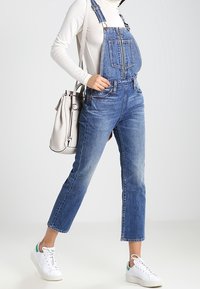 Denim overalls in medium blue with adjustable straps, front pocket detailing, and tapered legs, paired with white sneakers and a light bag.