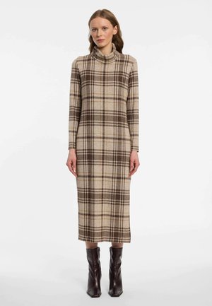 Frau trägt ein beige-braun kariertes Langarm-Rollkragenkleid mit dunkelbraunen Stiefeletten und steht vor einem weißen Hintergrund.