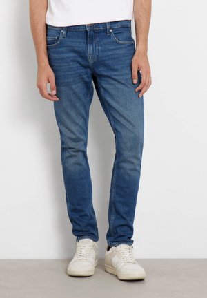 Jeans en denim bleu avec une coupe slim, présentant un design classique à cinq poches, un léger délavage et des détails de couture subtils. Associés à des baskets blanches.