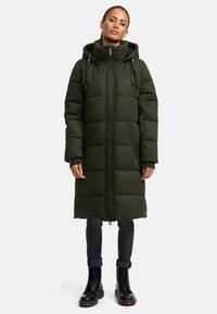 Polstret grøn parka med hætte, frontlynlås og snøre, sidelommer og knælang pasform, båret over sorte jeans og støvler.