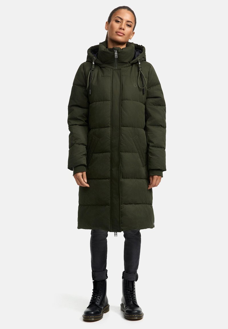 Polstret grøn parka med hætte, frontlynlås og snøre, sidelommer og knælang pasform, båret over sorte jeans og støvler.