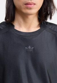 Melnā sporta T-kreklā ar apaļu kakla izgriezumu, kurā ir teksturēti plecu akcenti un reljefs logo uz krūtīm. Gluda auduma apdare.