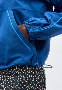 Χέρι ατόμου στην τσέπη ενός μπλε υφασμάτινου μπουφάν με ελαστικό τελείωμα και κορδόνι, που φοράει μαύρο παντελόνι με λευκά και μπλε σχέδια φύλλων.