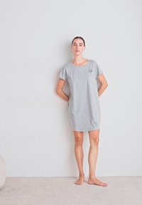 Šedé šaty s krátkým rukávem vyrobené z měkké látky, s uvolněným střihem a malou kapsou. Model stojí bos na neutrálním pozadí.
