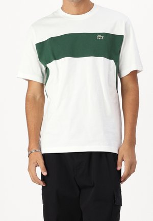 Mężczyzna w białym T-shircie z krótkim rękawem, zielonym poziomym panelem i małym logo na piersi, w połączeniu z czarnymi spodniami typu cargo.