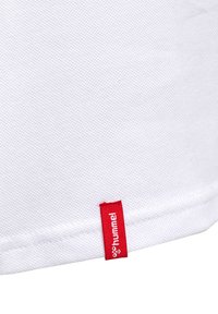 Witte gestructureerde stof met een klein rood rechthoekig label, genaaid vlakbij een naad, met witte tekst "hummel" en een logo.