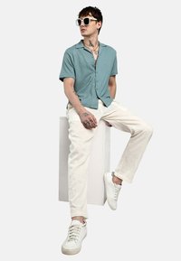 Camicia a maniche corte con bottoni in tessuto azzurro chiaro, abbinata a pantaloni beige chiaro e sneaker bianche. Gli accessori comprendono occhiali da sole.
