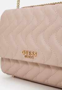 Roze handtas van nepleer met golvend patroon, gouden logo met "GUESS EST 1981" en kettingriem. Gladde textuur en gestructureerde vorm.