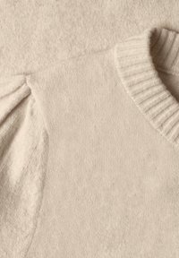Gros plan d'un pull en maille couleur crème montrant un col rond côtelé et une manche bouffante froncée sur un tissu à texture douce.