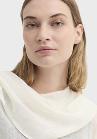 Écharpe en lin blanc drapée autour du cou, caractérisée par une texture douce et légère. Tissu lisse avec un léger éclat et un design minimaliste.