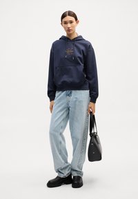 Sweatshirt azul-marinho com bolso frontal e logótipo dourado, combinado com jeans soltos azuis claros e sapatos pretos. O modelo segura uma bolsa preta.