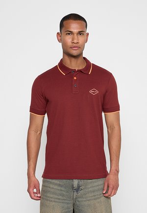 Mann trägt ein kastanienbraunes Polohemd mit orangefarbener Einfassung und Logo auf der Brust, kombiniert mit hellgrauen Jeans, steht vor einem einfarbigen Hintergrund.